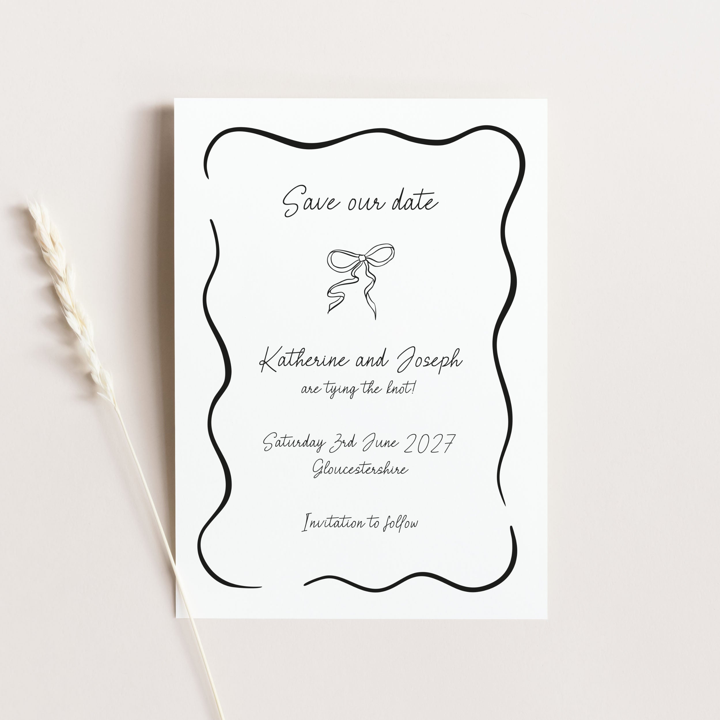A white 'Save our date' card with a black wavy border and elegant script text, photographed on a light beige background with a dried wheat stem to the left.