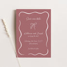 A dusty rose 'Save Our Date' card with white script text and a white wavy border, styled flat-lay on a light beige background with a dried wheat stalk.