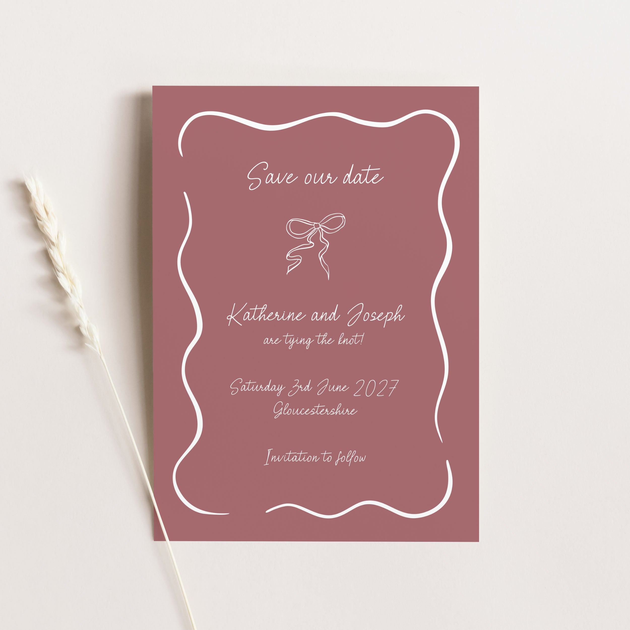 A dusty rose 'Save Our Date' card with white script text and a white wavy border, styled flat-lay on a light beige background with a dried wheat stalk.