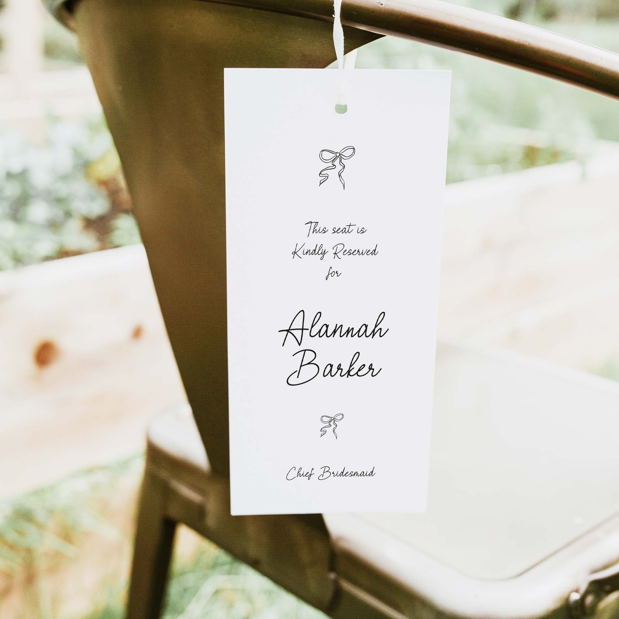 A white printed reserved seat card hanging from a metal chair outdoors. The card has black script text reading 'This seat is kindly reserved for Alannah Barker' and 'Chef Bridesmaid' with small bow illustrations.