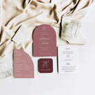 A coordinated wedding invitation suite arranged on a white surface with draped champagne silk. The set includes a rose pink arch invitation, a matching rounded arch detail card, a white RSVP card, and a small dark burgundy square card, styled with clear textured glassware.