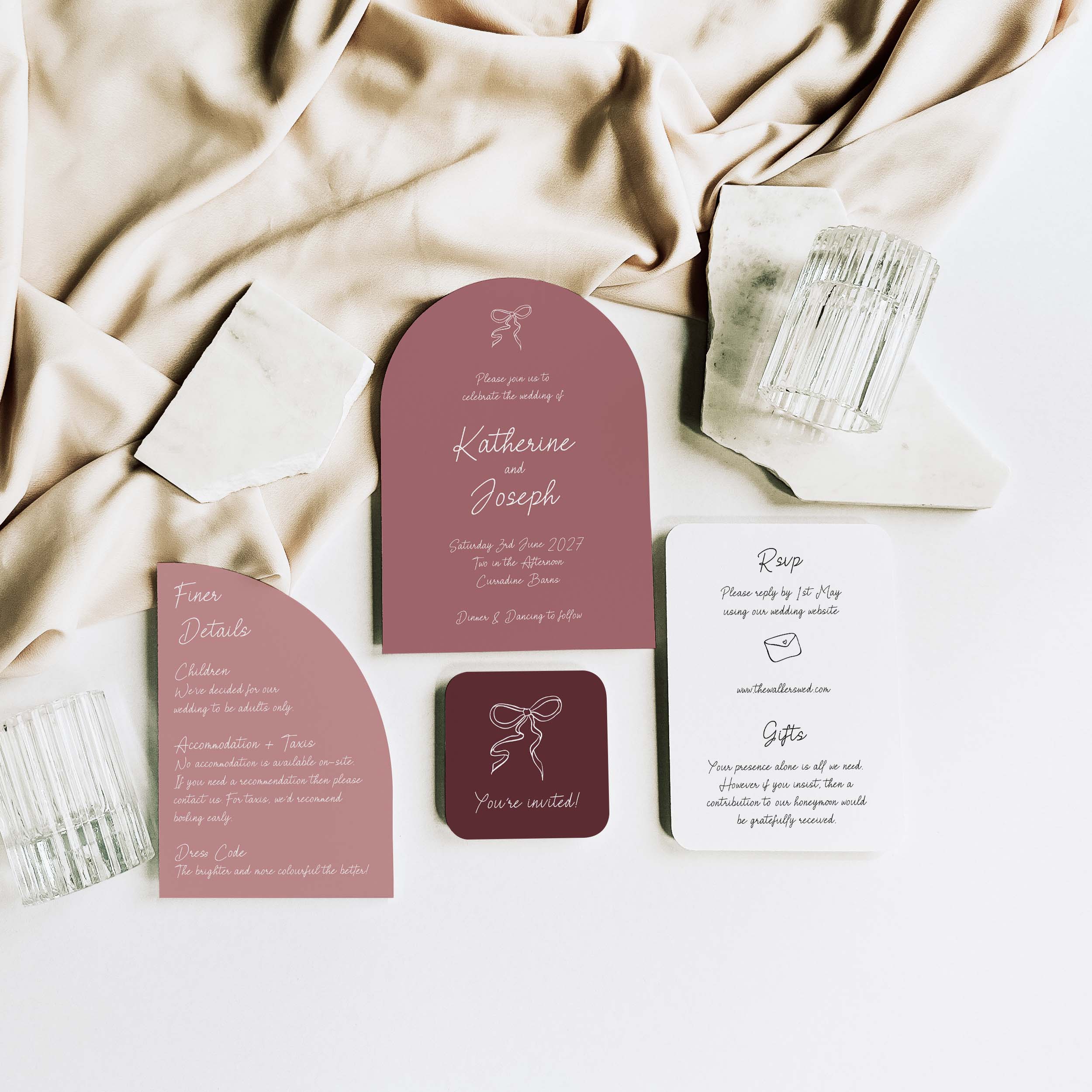 A coordinated wedding invitation suite arranged on a white surface with draped champagne silk. The set includes a rose pink arch invitation, a matching rounded arch detail card, a white RSVP card, and a small dark burgundy square card, styled with clear textured glassware.