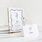 A tabletop wedding stationery set featuring a vertical table number frame with a metallic rose gold frame and a matching white place card with a black bow illustration and the name 'Hattie'.