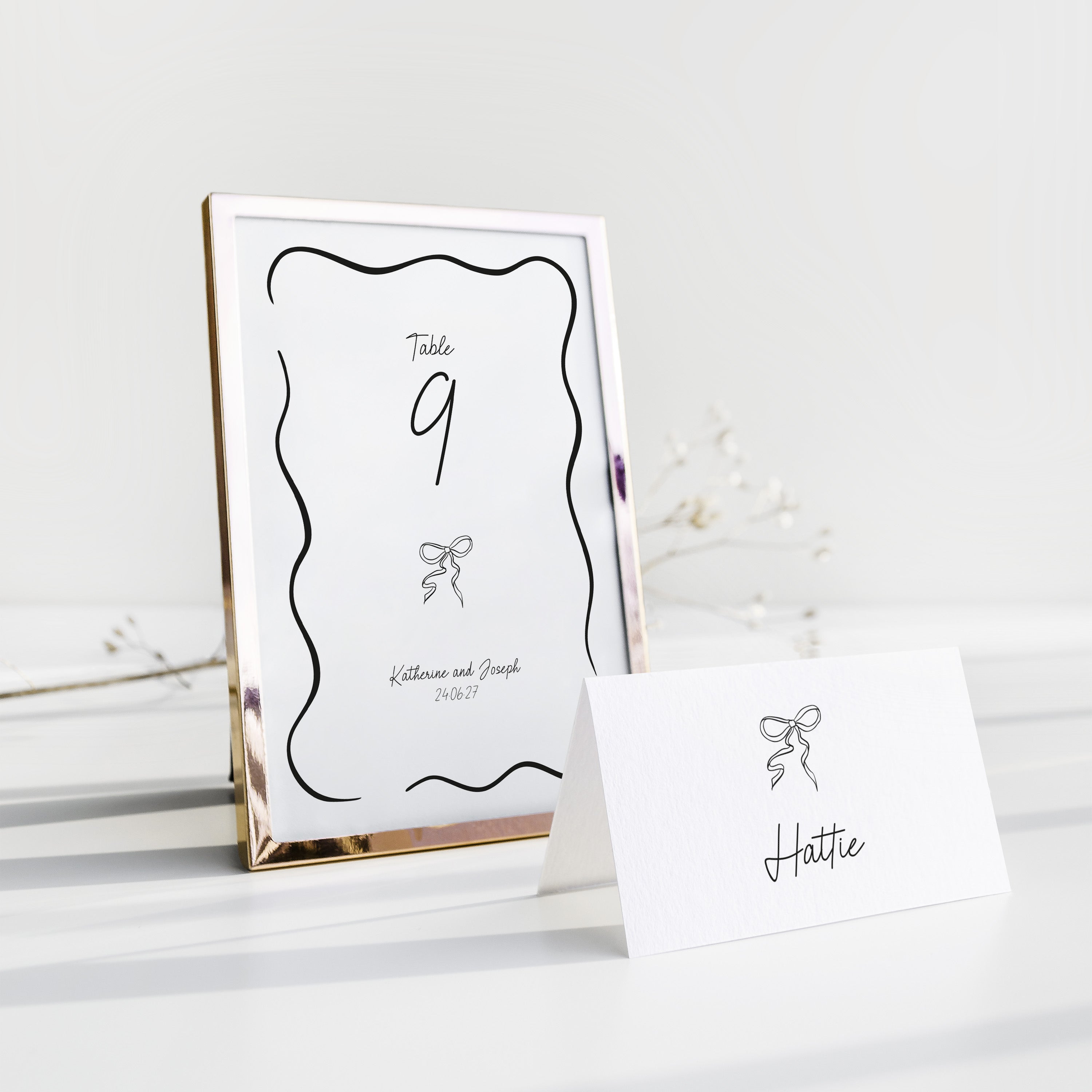 A tabletop wedding stationery set featuring a vertical table number frame with a metallic rose gold frame and a matching white place card with a black bow illustration and the name 'Hattie'.