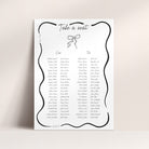 A white wedding seating chart print with a black decorative border, the heading 'Take a seat,' and printed names arranged in columns under labeled sections 'One' and 'Two,' shown against a light beige wall.