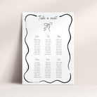 A wedding seating chart print with a decorative black wavy border, the heading 'Take a seat,' and tables for One, Two, Three, Four, Five, and Six printed in black text on a white sheet, shown against a light beige wall.
