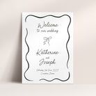A vertical white wedding welcome sign with a black scalloped border and elegant script text reading 'Welcome to our wedding' and 'Katherine and Joseph,' shown leaning against a light beige wall.
