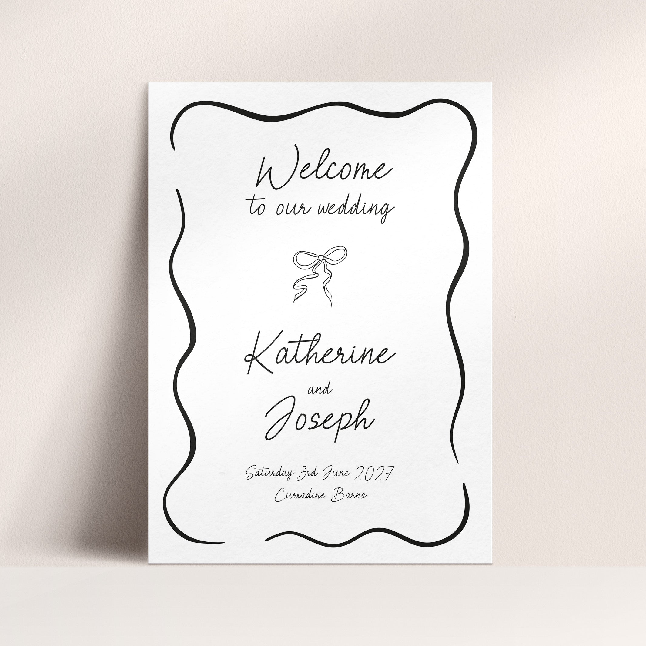 A vertical white wedding welcome sign with a black scalloped border and elegant script text reading 'Welcome to our wedding' and 'Katherine and Joseph,' shown leaning against a light beige wall.