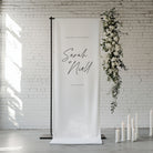 Wedding backdrop with floral arrangement and hanging linen welcome sign against a white brick wall.