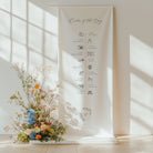 Floral arrangement in a pot on a light wooden floor with a white wall and a decorative banner.