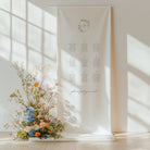 Floral arrangement in a vase next to a seating chart on a white wall with sunlight filtering through.