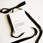 A white save the date card for a wedding shown above a white background and alongside a black satin ribbon tied into a bow