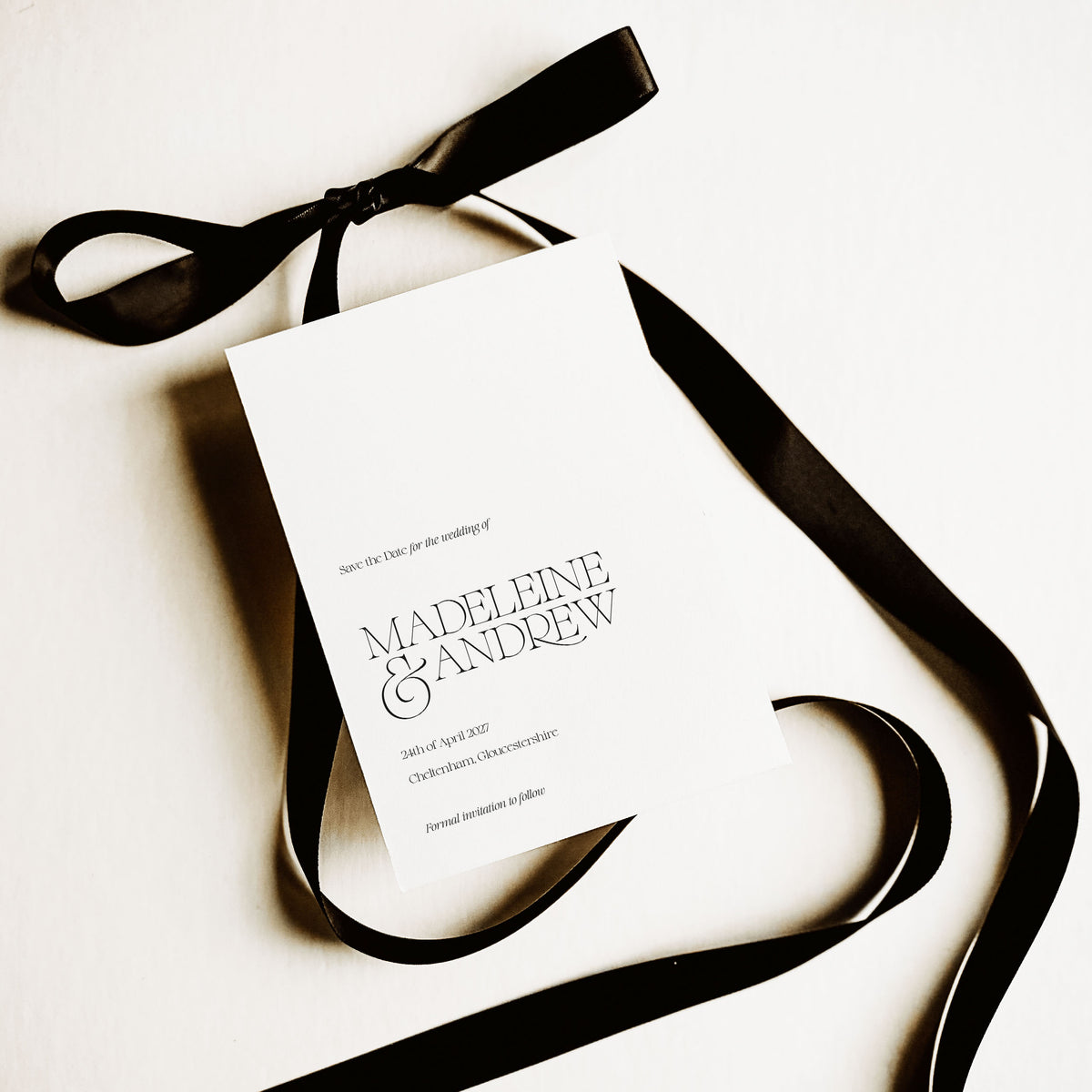 A white save the date card for a wedding shown above a white background and alongside a black satin ribbon tied into a bow