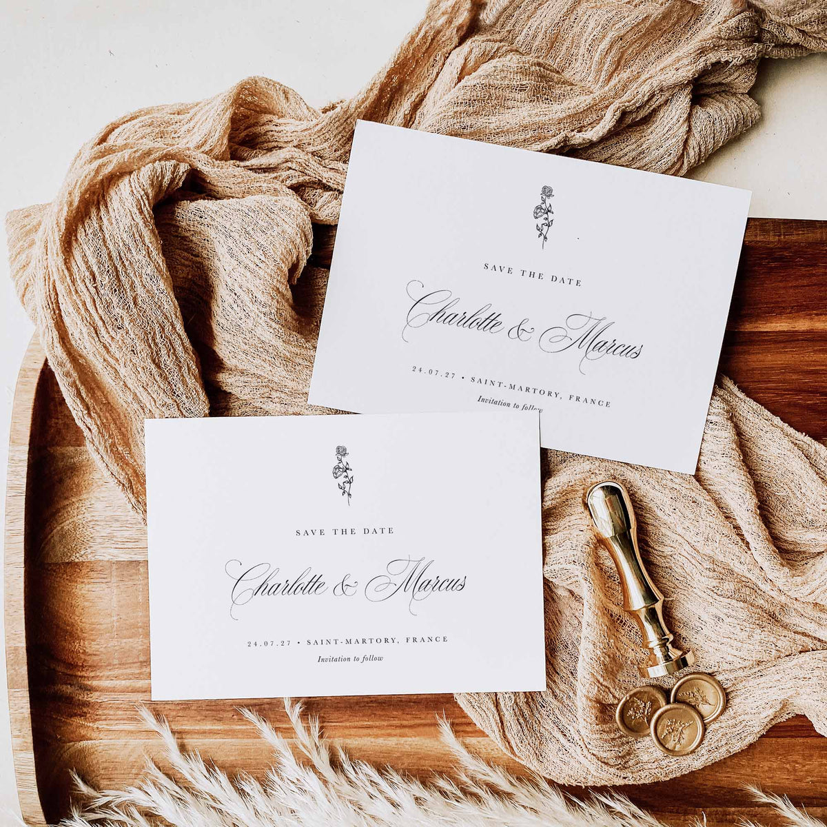 Two white save the date cards in landscape orientation displayed above neutral coloured fabric, wooden display board and alongside pampass grass, wax stamp and wax seals
