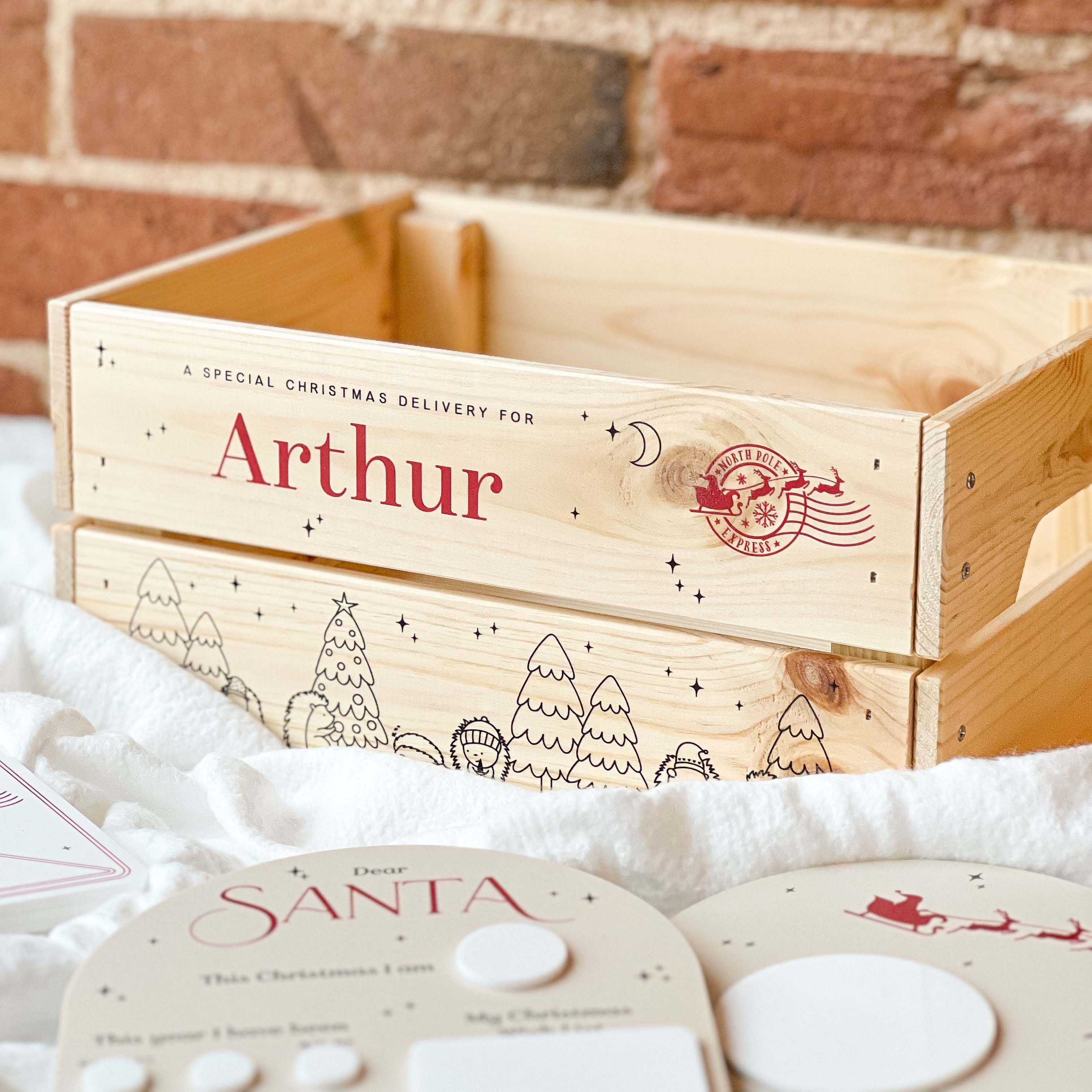 Wooden Christmas Eve gift box with 'Arthur' on it, surrounded by festive items against a brick wall background.