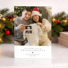 Christmas-themed pregnancy announcement card with a couple holding a baby onesie and ultrasound photo.