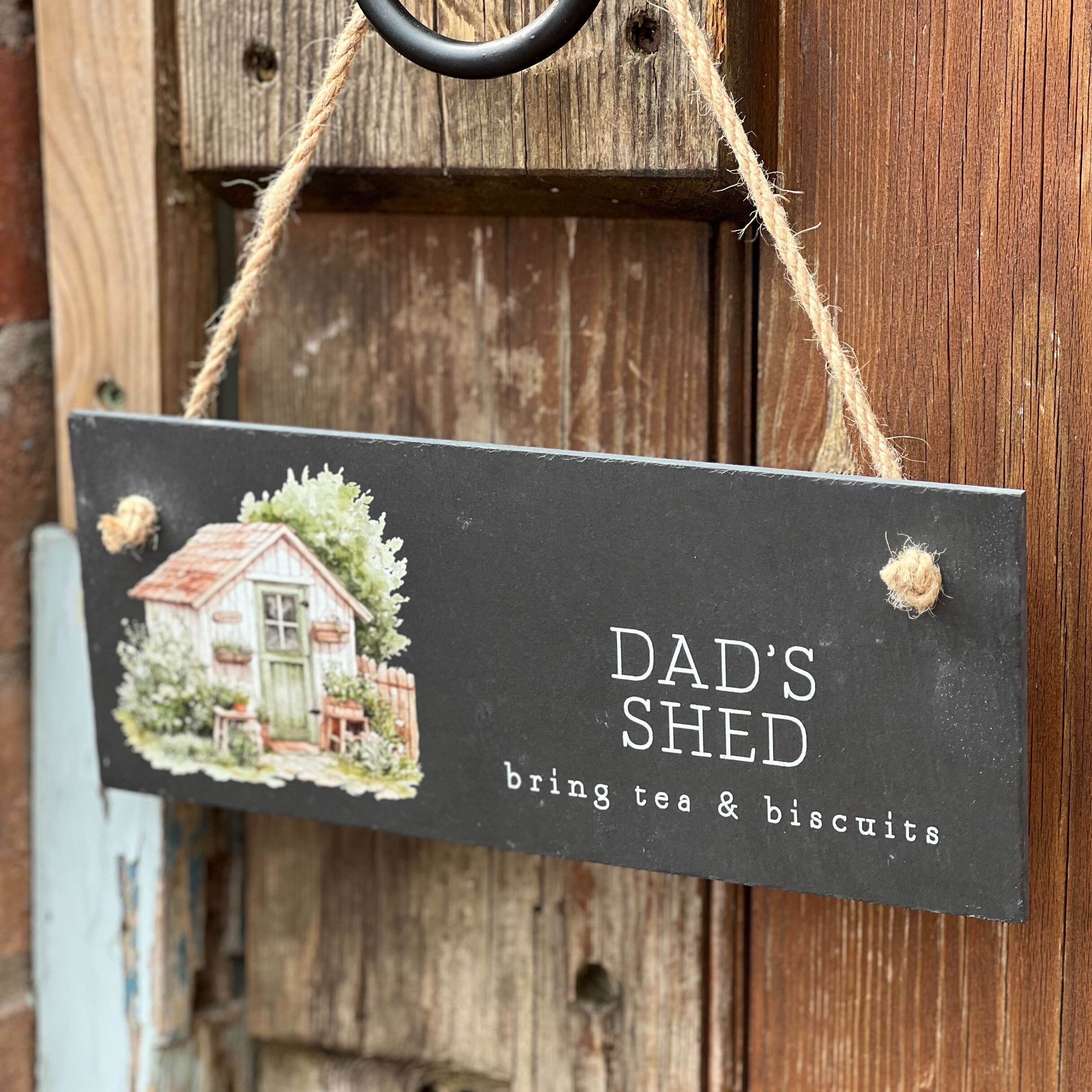 Decorative sign with a house illustration and text 'Dad's Shed bring tea & biscuits' hanging on a wooden door.