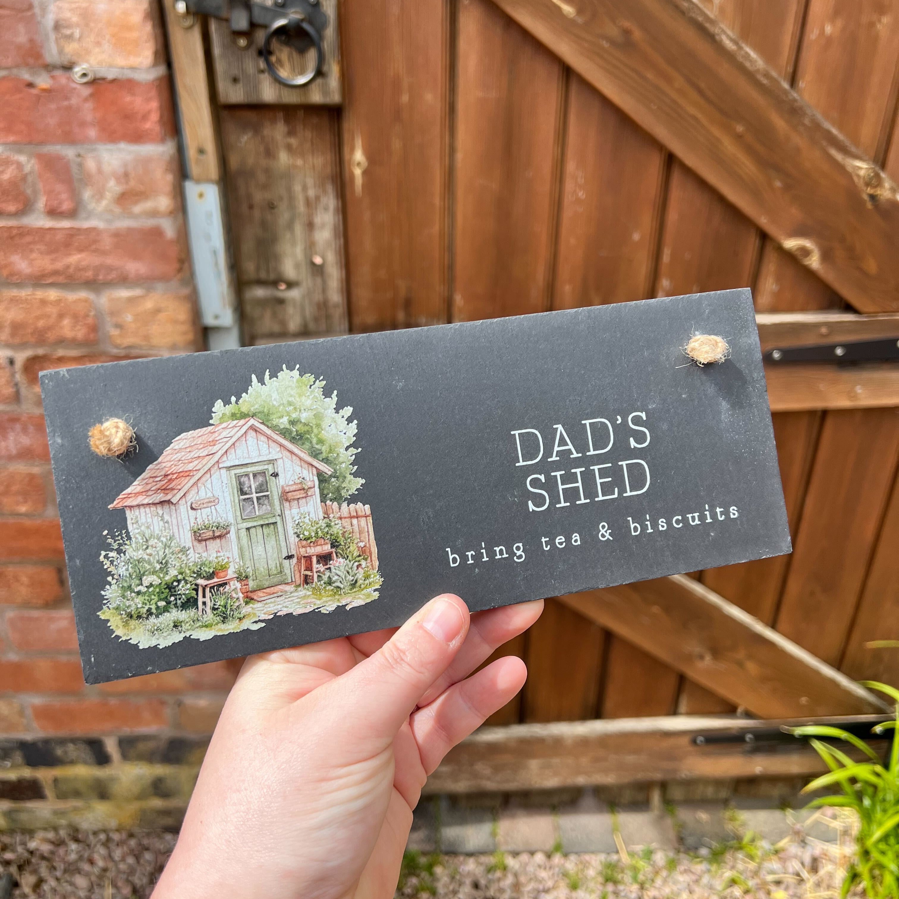Hand holding a sign with 'Dad's Shed' text and illustration in front of a wooden gate.