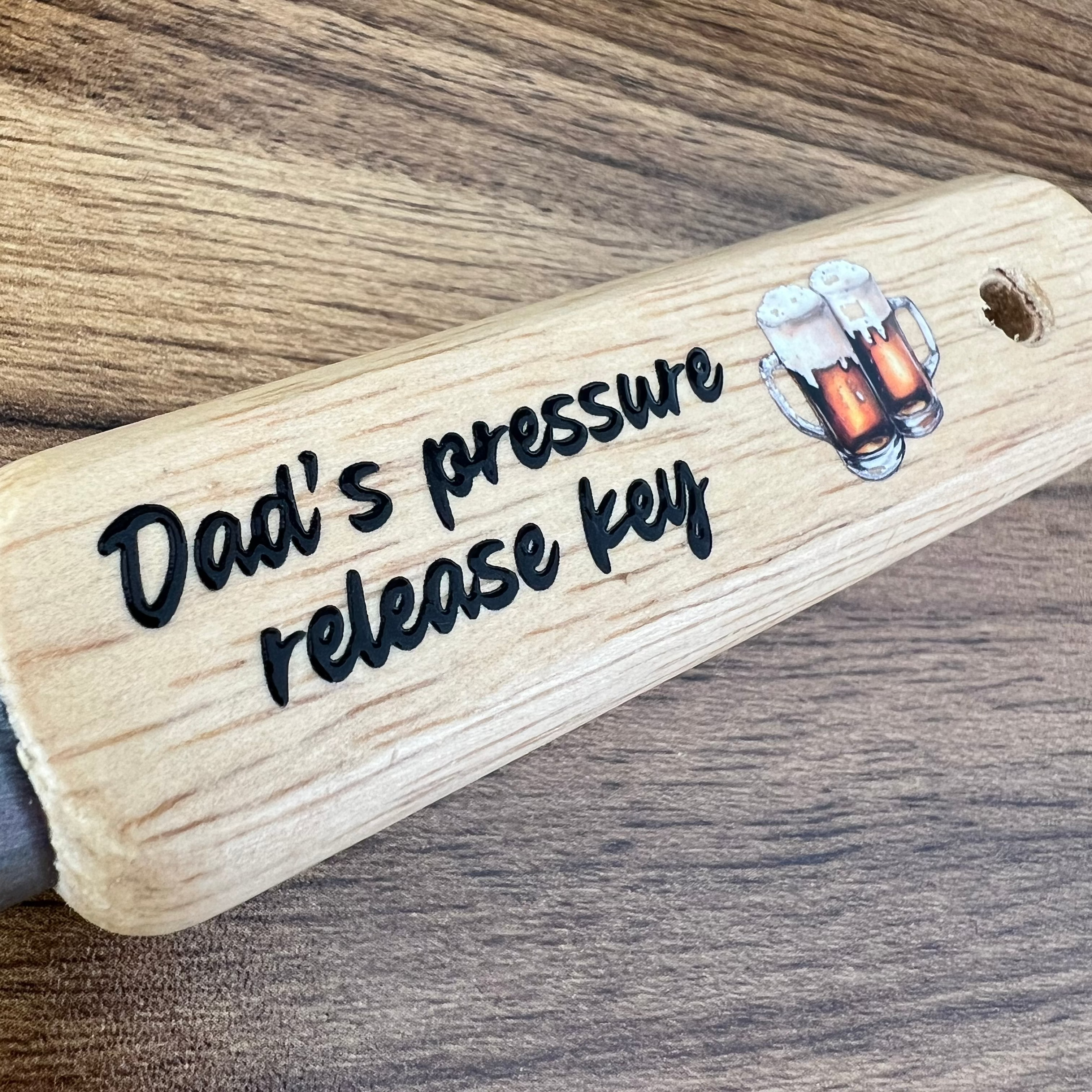 Wooden bottle opener with 'Dad's pressure release key' text and beer graphics on a wooden surface