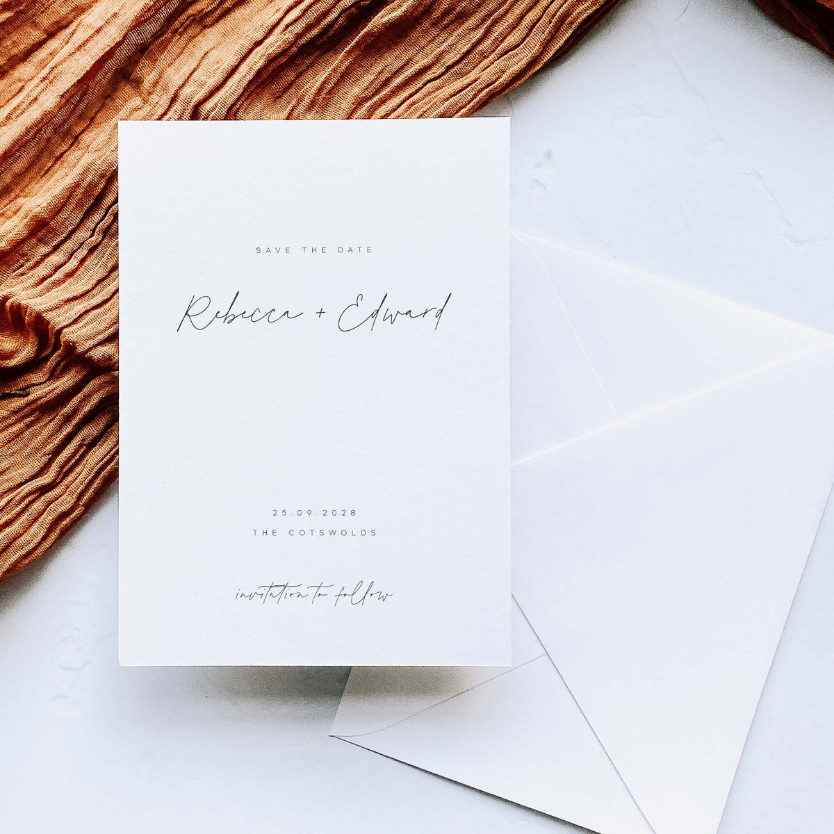 A white save the date card for a wedding with modern text, displayed with a white diamond flap envelope on a white background alongside copper coloured fabric