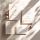 A coordinated wedding invitation suite arranged on a warm cream fabric background with soft diagonal shadows. The set includes a main invitation, an acceptance card, a marriage proposal card, a program/booklet, and small pink flowers used as styling props.
