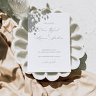 White save the date card with elegant handwritten text, displayed on a white seashell style plate above neutral coloured fabric and alongside a glass bottle
