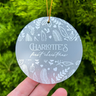 A round frosted glass Christmas ornament with white botanical illustrations and the personalized text 'Charlotte's first christmas', held in a hand against a background of green pine branches.