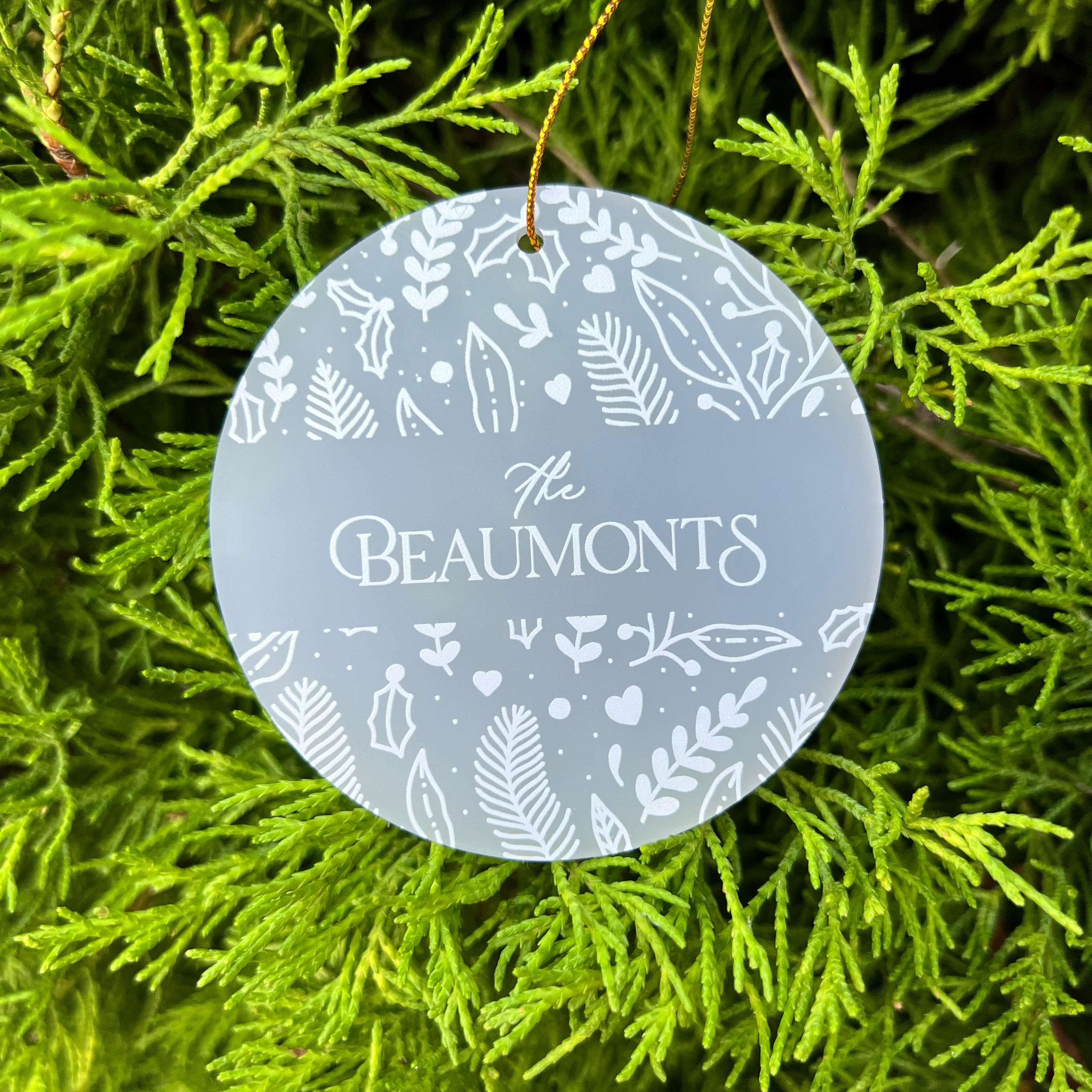 A round frosted glass Christmas ornament hanging from a gold string, featuring white botanical illustrations and the text 'The Beaumonts' in the center, set against a background of green pine branches.