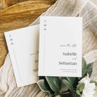 Two white save the date cards for a wedding displayed on sheer white fabric and on a wooden display board, alongside green and white foliage.