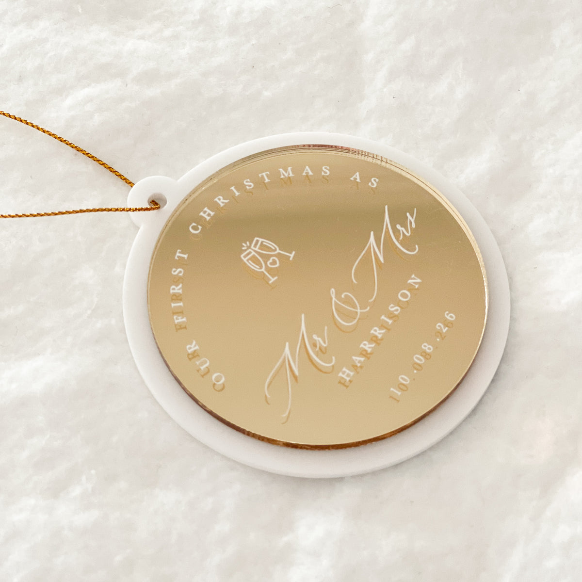 A personalized round Christmas ornament with a gold center and white outer rim, hanging from a thin gold string. The ornament is engraved with 'OUR FIRST CHRISTMAS AS Mr & Mrs HARRISON' and the date '10.