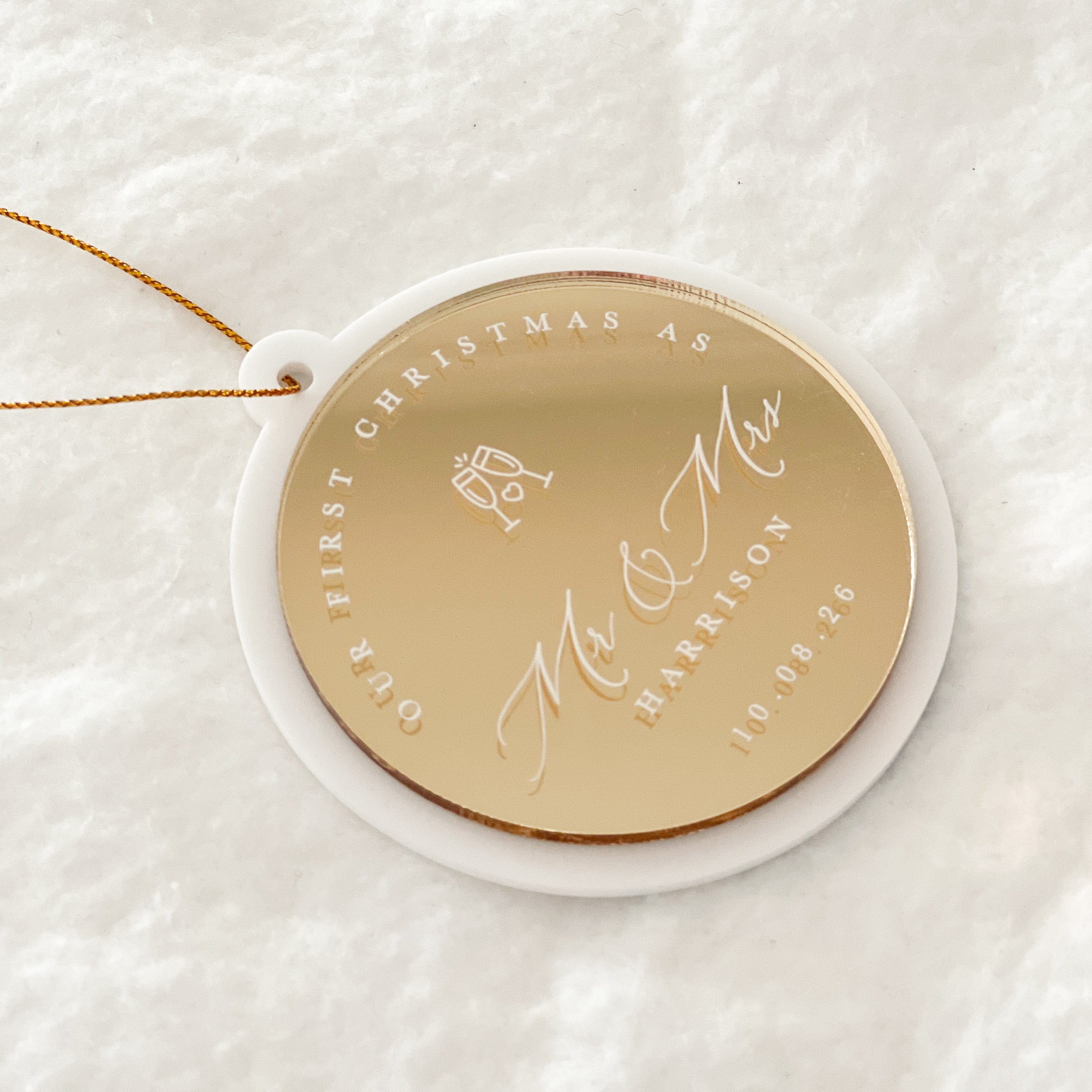 A personalized round Christmas ornament with a gold center and white outer rim, hanging from a thin gold string. The ornament is engraved with 'OUR FIRST CHRISTMAS AS Mr & Mrs HARRISON' and the date '10.