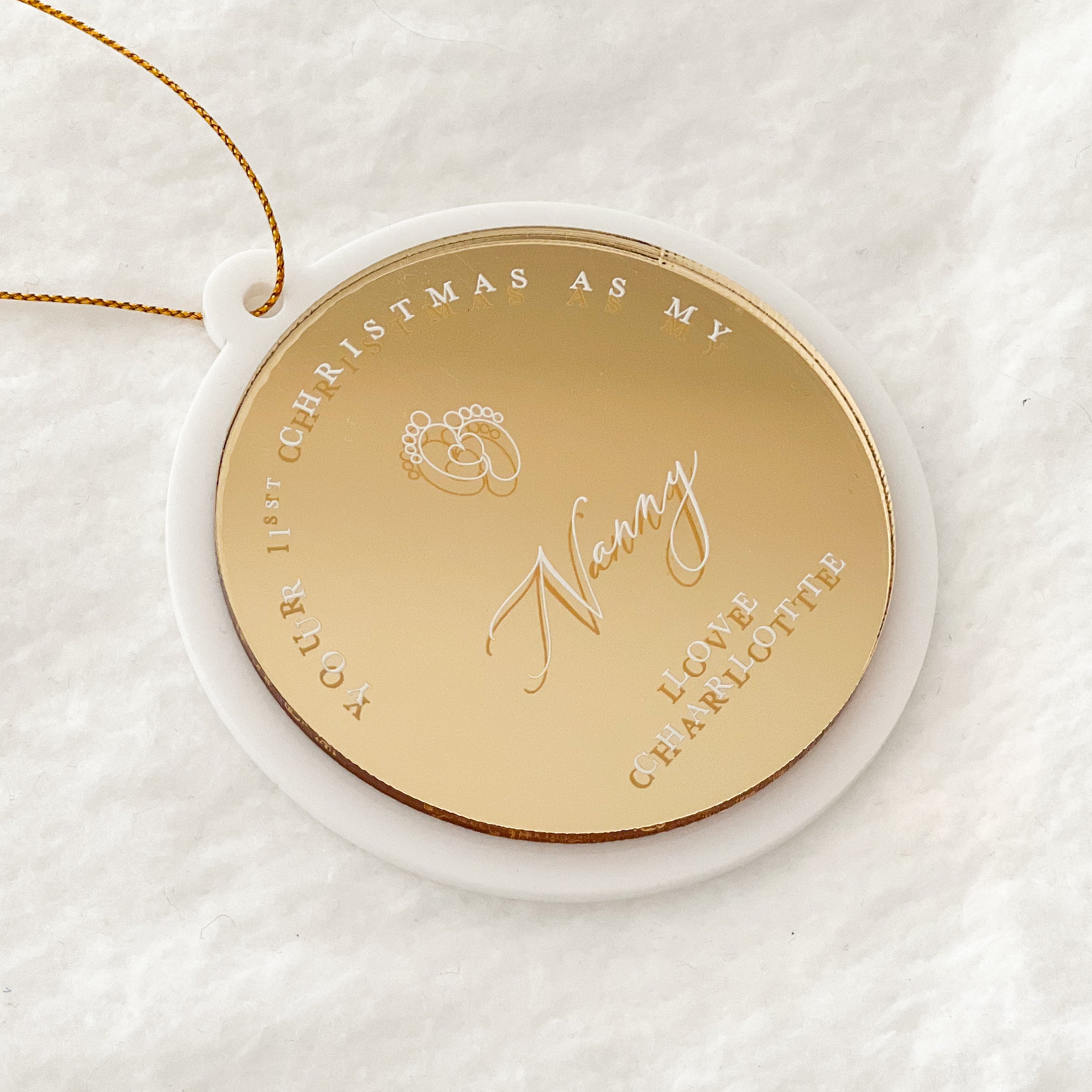 A personalized round gold Christmas ornament with a white backing ring and a gold hanging string, featuring engraved text and wedding rings, displayed on a soft white textured background.