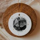 A round white personalized Christmas ornament with a gold hanging string, featuring a black-and-white photo of a grandfather and grandmother with the text '1st Christmas as my grandpa' and 'Love Noah,' displayed on a round wooden board.