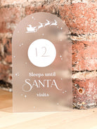 A frosted acrylic Christmas advent countdown sign with white laser-cut designs of Santa Claus, reindeer, and stars. The sign is displayed on a light wooden surface with a rustic red brick wall in the background.