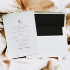 A white wedding invitation card with elegant black typography and monogram lettering, placed on top of a matching white envelope with a black insert, styled on a soft beige fabric background with decorative dried grass.