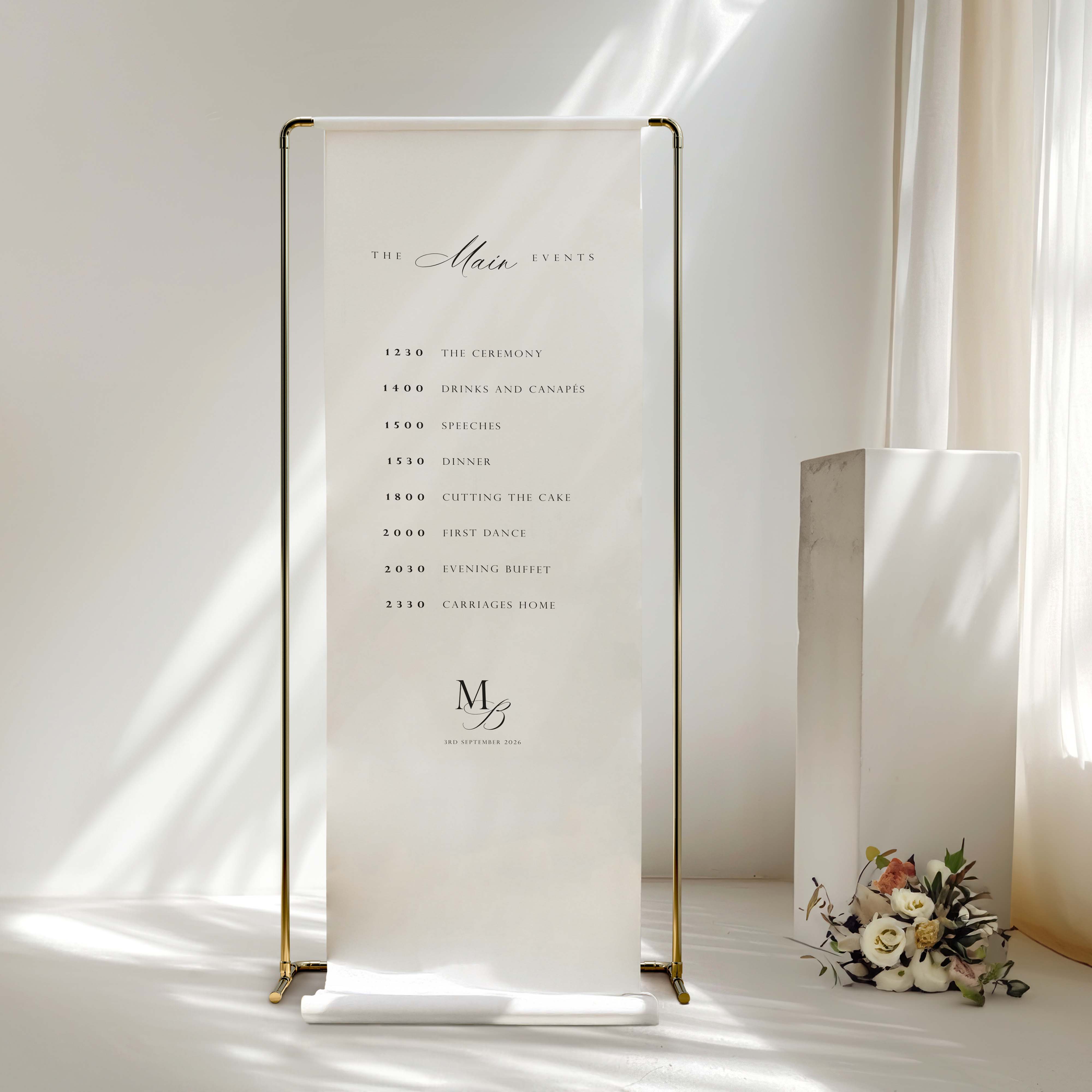 A vertical white wedding reception seating chart or program stand with gold metal framing, displayed on a white floor against a softly lit cream wall. The printed text includes 'The Main Events' and a timeline, with a monogram logo near the bottom.