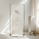 A fabric wedding welcome sign hanging from a gold frame in front of a window.
