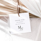 A white square thank-you tag with black text reading 'THANK YOU FOR SHARING THIS DAY WITH US' and a monogram, hanging from a brown string over dried palm leaves and white fabric.