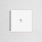 A white spiral-bound wedding guest book with a black wire binding and a minimalist black monogram logo on the cover, displayed on a light grey textured fabric background.