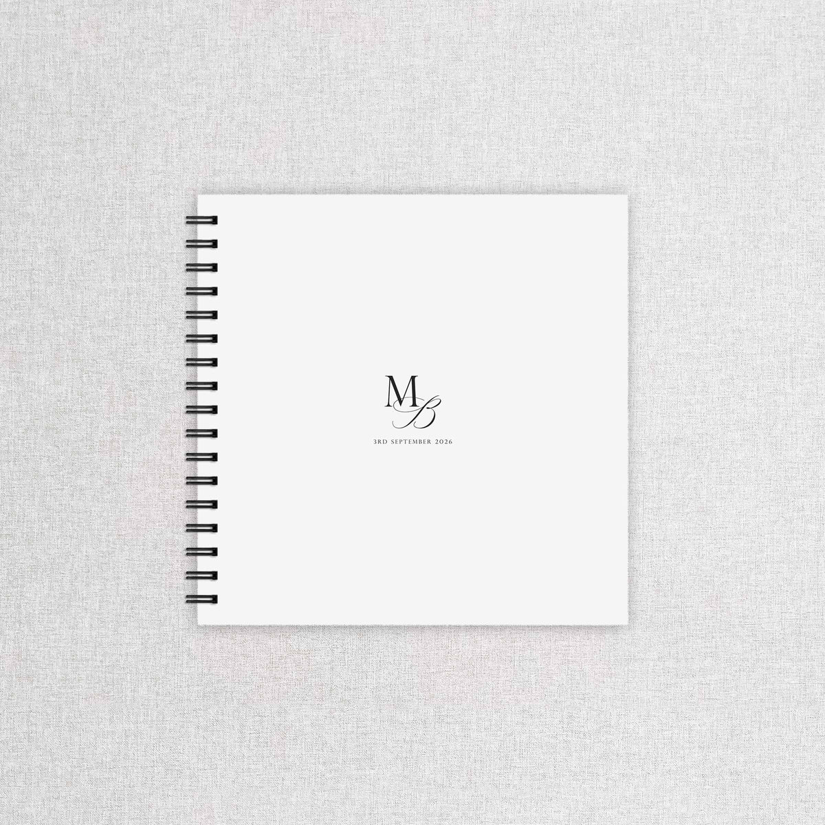 A white spiral-bound wedding guest book with a black wire binding and a minimalist black monogram logo on the cover, displayed on a light grey textured fabric background.