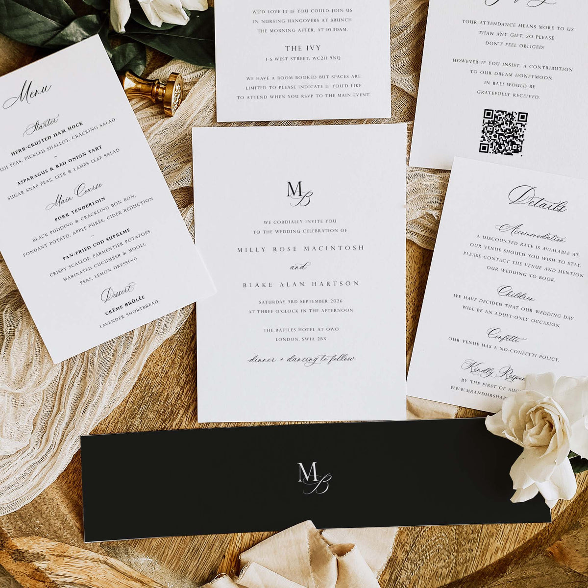 A styled flat-lay of wedding stationery, including printed invitation cards, a QR code, and a black envelope with a white monogram logo, arranged on textured beige fabric and wood with a cream rose.
