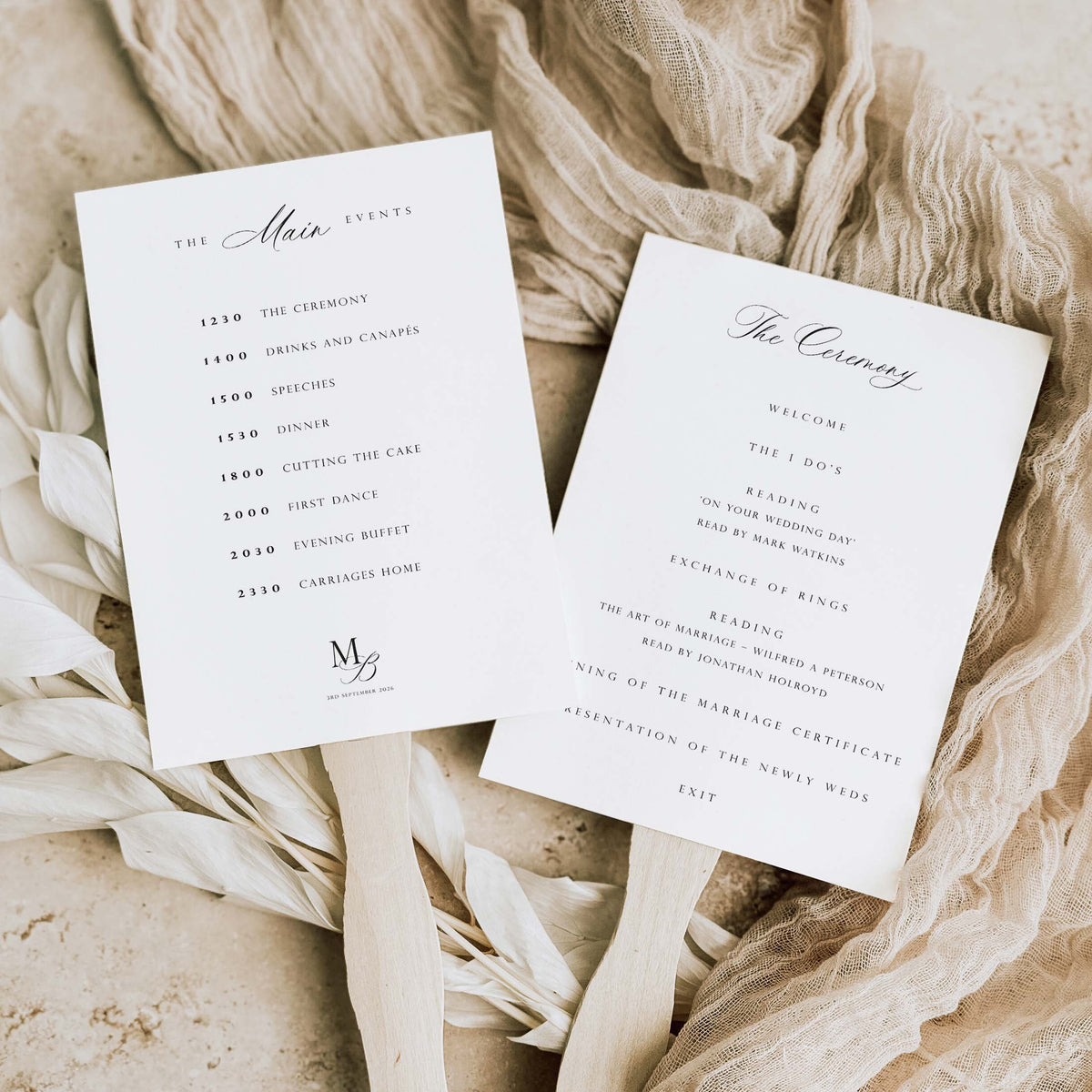 Two white wedding program cards with elegant black script and serif text, displayed on wooden sticks over a neutral beige fabric and dried floral arrangement.