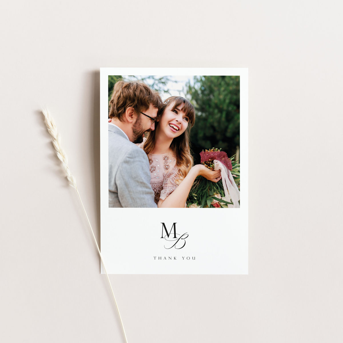 A vertical white thank-you card with a printed photo of a smiling couple at the top and a large monogram 'MB' with 'THANK YOU' below, styled on a light beige background with a dried wheat stem to the left.