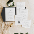 A styled flat-lay of wedding stationery on a light beige textured surface with dried flowers and small gold bells. The set includes a black pocketfold with a white invitation card, a guest information card, a wedding program, a guest listing card, and a morning-after card.
