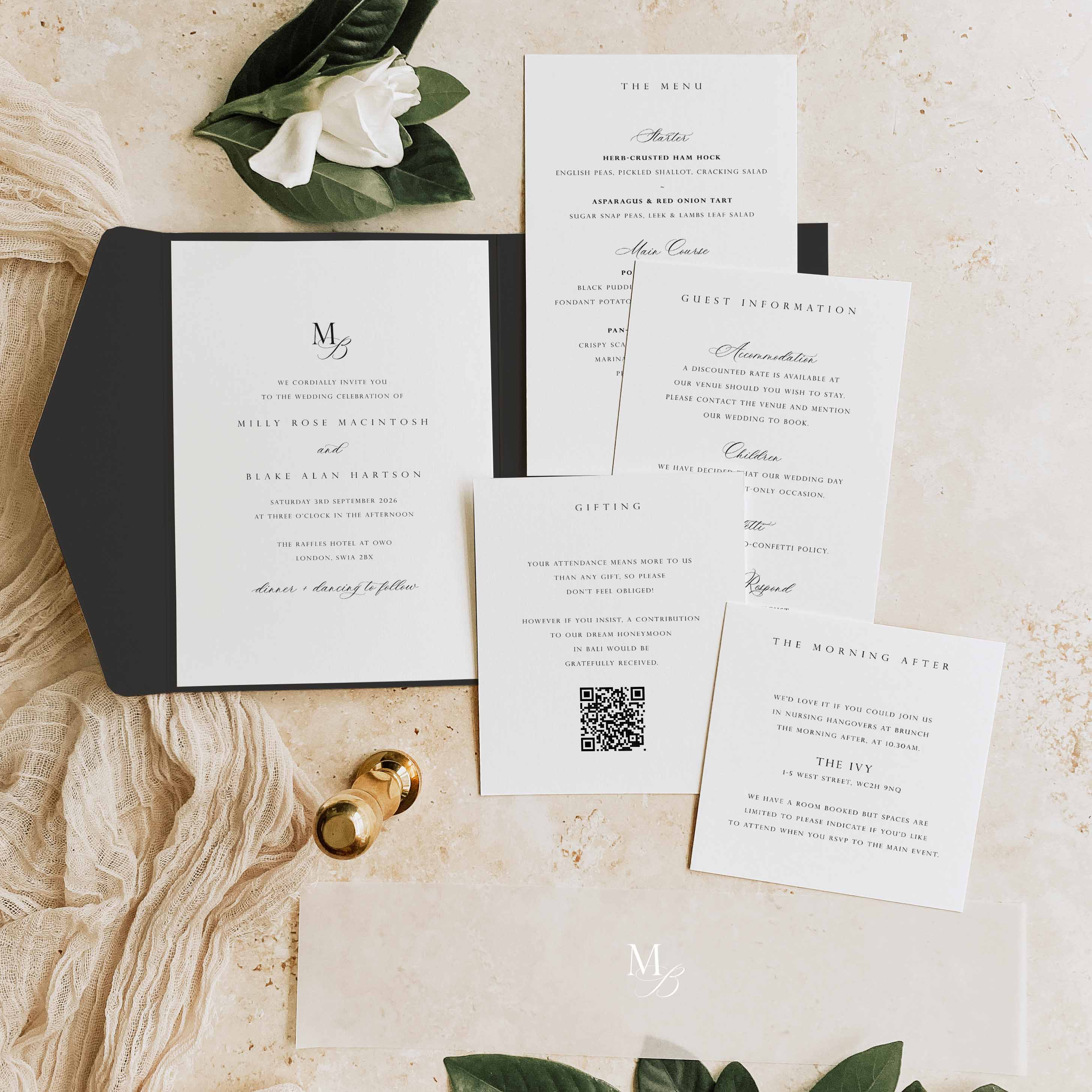 A styled flat-lay of wedding stationery on a light beige textured surface with dried flowers and small gold bells. The set includes a black pocketfold with a white invitation card, a guest information card, a wedding program, a guest listing card, and a morning-after card.