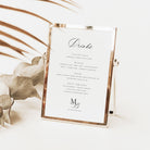 A small rectangular tabletop drink menu sign with a white card and metallic rose-gold frame, styled on a light surface with dried leaves in the background.