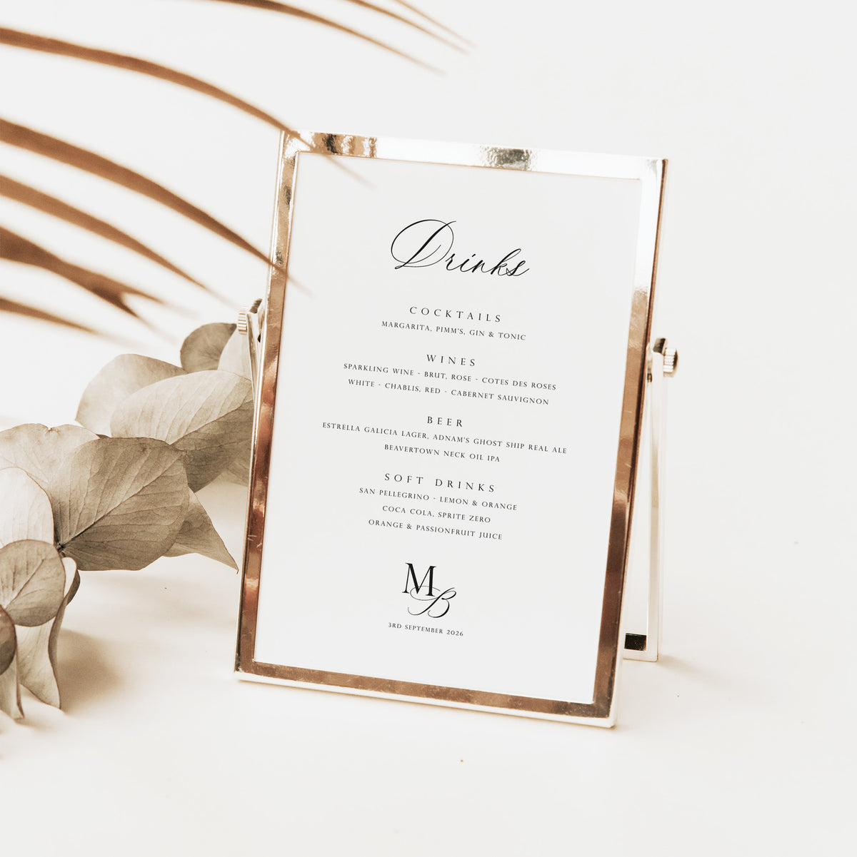 A small rectangular tabletop drink menu sign with a white card and metallic rose-gold frame, styled on a light surface with dried leaves in the background.