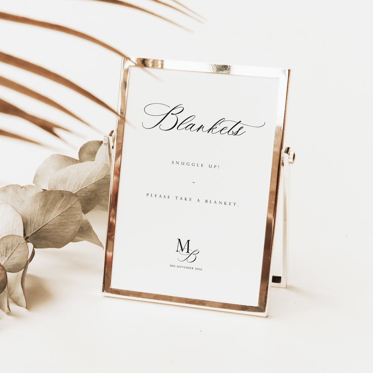 A rose gold metal picture frame with a white insert sign that reads 'Blankets' and 'Please Take a Blanket,' styled on a white surface with dried leaves and palm leaves in the background.