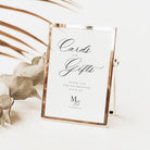 A rectangular gold-framed sign with a white insert stands on a light surface. The insert reads 'Cards and Gifts' in elegant script, with smaller thank-you text below and a monogram logo.