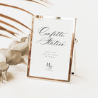 A rectangular freestanding sign with a metallic rose-gold frame and a white insert reading 'Confetti Station' in black script, styled on a light surface with dried leaves and palm fronds.